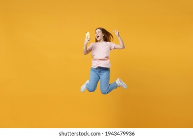 Full length of young expressive friendly overjoyed woman 20s in basic pastel pink t-shirt jump high holding mobile cell phone show victory v-sign gesture isolated on yellow background studio portrait