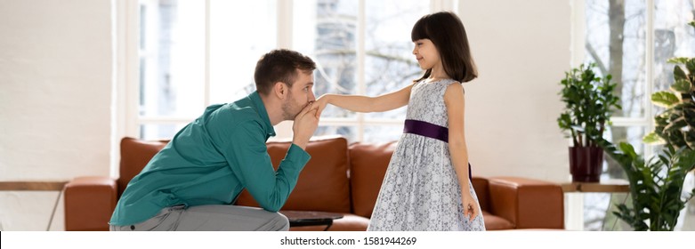 Full Length Young Father Standing On Knee, Kissing Hand Of Smiling Little Daughter, Looking At Eyes, Inviting For Dance. Joyful Small School Girl Ready Training Performance On Modern Living Room.