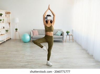 Full length of young fit Indian lady in VR headset doing virtual yoga, standing in tree pose at home, copy space. Athletic Eastern lady using modern technologies for domestic workout - Powered by Shutterstock