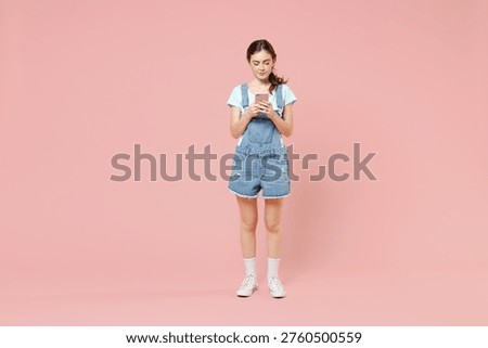 Full length young friendly smiling happy woman wearing trendy denim clothes blue t-shirt hold in hand use mobile cell phone isolated on pastel pink background studio portrait. People lifestyle concept