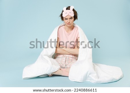 Full length young frowning woman in pajamas jam sleep eye mask rest at home sit wrap covered blanket duvet hold hands crossed folded isolated on pastel blue background Bad mood night bedtime concept