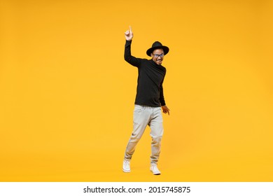 Full length young fun african man 20s wearing stylish black hat shirt eyeglasses doing winner gesture pointing index finger up walk going isolated on yellow orange color background studio portrait.