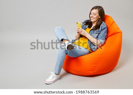 Full length young fun caucasian woman wear denim jacket yellow t-shirt use mobile cell phone browsing sitting in bean bag chair isolated on grey background studio portrait. People lifestyle concept.