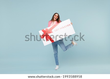 Full length young fun excited woman 20s wearing pastel pink clothes glasses hold large huge gift voucher flyer mock up with raised up leg isolated on blue background studio People lifestyle concept