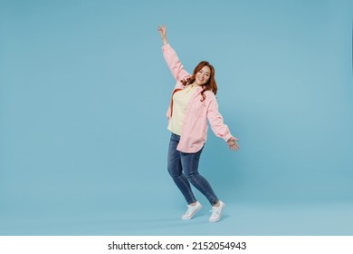 Full length young fun joyful smiling redhead chubby overweight woman 30s wearing in pink shirt jeans leaning back stand on toes with oustretched hands hands dance isolated on pastel blue background