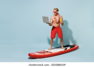 Full length young fun man in red shorts swimsuit hawaii lei glasses stand on sup board hold alcohol cocktail laptop pc computer isolated on pastel blue background Summer vacation sea sun tan concept.