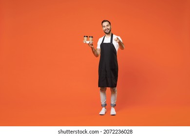 Full length young fun man barista barman employee in apron white t-shirt work in coffee shop hold paper takeaway delivery cups point finger on you isolated on orange background Small business startup