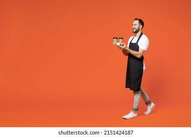 Full length young fun man 20s barista barman employee in black apron white t-shirt work in coffee shop hold paper takeaway delivery cups walk go isolated on orange background Small business startup - Powered by Shutterstock