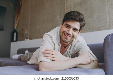 Full length young fun smiling cheerful happy man in casual clothes beige shirt pink t-shirt prop up forehead look camera lying on grey sofa rest indoors at home on weekends. People leisure concept.