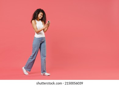 Full Length Young Fun Smiling Happy Satisfied Positive African American Woman 20s In Casual White Tank Shirt Hold In Hand Using Mobile Cell Phone Chat Online Walking Going Isolated On Pink Background.