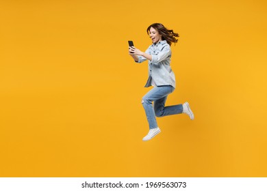 Full length of young fun sporty student woman 20s wear casual stylish denim shirt white t-shirt run jump high hold mobile cell phone use fast internet isolated on yellow background studio portrait.