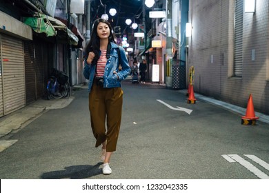 Full Length Of Young Girl In Brown Trousers Walking By Path In City. Businesswoman After Work Going Back To Home At Night. Quiet Street With Traffic Cones Beside The Houses.