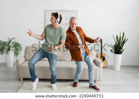 full length of young guy with senior dad in wireless headphones listening music and dancing in living room