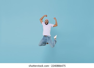 Full length young happy cool overjoyed man of African American ethnicity wear violet t-shirt hat do winner gesture jump high up clench fist celebrate isolated on pastel blue background studio portrait
