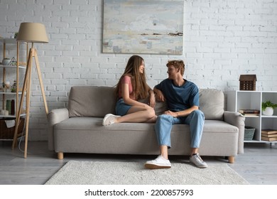 Full Length Of Young And Happy Couple Sitting On Couch And Looking At Each Other In Living Room