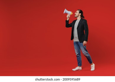 Full length young happy expressive fun businessman latin man 20s wearing black striped jacket grey shirt scream in megaphone announces discounts sale hurry up isolated on red color background studio.