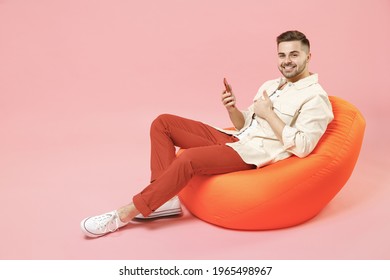 Full Length Young Happy Fun Man In Jacket White T-shirt Sitting In Bean Bag Chair Using Mobile Cell Phone Chat Online In Social Network Show Thumb Up Gesture Isolated On Pastel Pink Background Studio.