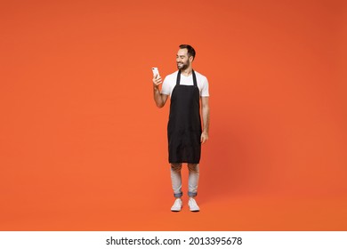 Full Length Young Happy Fun Man Barista Bartender Barman Employee In Black Apron White T-shirt Work In Coffee Shop Using Mobile Cell Phone Isolated On Orange Background. Small Business Startup Concept