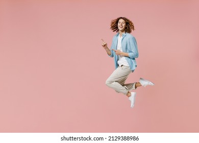 Full Length Young Happy Fun Cool Man 20s With Long Curly Hair Wear Blue Shirt White T-shirt Point Index Finger Aside On Workspace Area Jump High Isolated On Pastel Plain Pink Wall Background Studio.