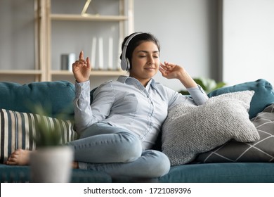 Full length young happy indian girl resting on cozy sofa, listening to energetic music in headphones. Smiling peaceful carefree young woman enjoying spending free time with favorite audio tracks.