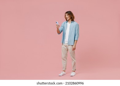 Full Length Young Happy Satisfied Cool Man With Long Curly Hair Wear Blue Shirt White T-shirt Hold In Hand Use Mobile Cell Phone Isolated On Pastel Plain Pink Color Wall Background Studio Portrait.