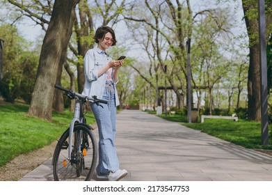 Full length young happy woman in jeans clothes stand near bicycle bike on sidewalk in city spring park outdoors use mobile cell phone chat online. People active urban healthy lifestyle cycling concept