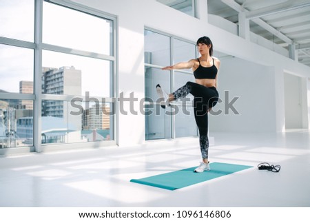 Full length of a young healthy woman doing stretching exercise on the mat at gym. Female in sportswear standing on one leg and touching the toe of the outstretched leg with hand.