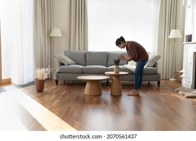 Full length of young Indian ethnicity housewife standing in modern spacious living room create cosiness at home, arranging flowers bunch in vase. Housework routine, interior designer workflow concept