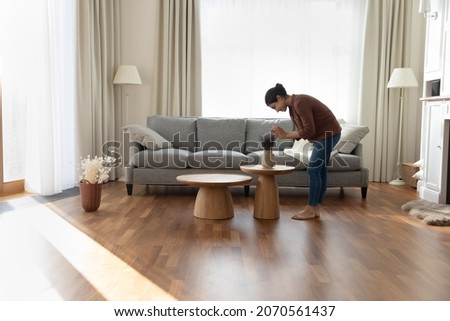 Full length of young Indian ethnicity housewife standing in modern spacious living room create cosiness at home, arranging flowers bunch in vase. Housework routine, interior designer workflow concept