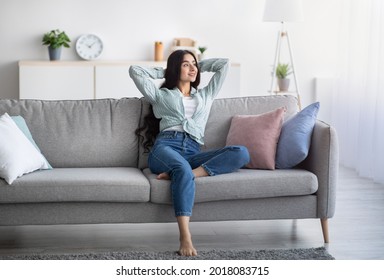 Full Length Of Young Indian Lady Relaxing On Lazy Weekend At Home, Sitting On Couch With Hands Behind Her Head, Copy Space. Young Woman Having Day Off, Chilling On Comfy Sofa Indoors