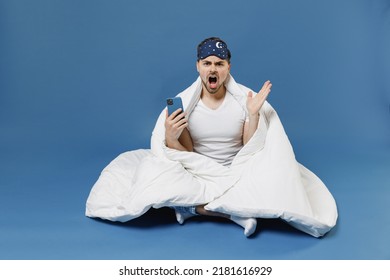 Full length young indignant man in pajamas jam sleep mask rest relax home sit wrap under cover blanket duvet use mobile cell phone spread hands isolated on dark blue background Night bedtime concept