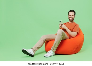 Full Length Young Man 20s Wear Casual Orange T-shirt Sit In Bag Chair Hold In Hand Use Mobile Cell Phone Isolated On Plain Pastel Light Green Color Background Studio Portrait. People Lifestyle Concept