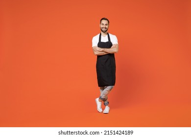 Full length young man barista bartender barman employee in black apron white t-shirt work in coffee shop hold hands crossed folded isolated on orange background studio. Small business startup concept