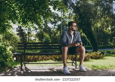 Full length young man in blue shirt glasses sit on bench use mobile cell phone chat look aside rest relax in spring green city park sunshine lawn outdoors on nature Urban lifestyle leisure concept.