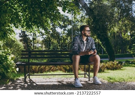 Full length young man in blue shirt glasses sit on bench use mobile cell phone chat look aside rest relax in spring green city park sunshine lawn outdoors on nature Urban lifestyle leisure concept.
