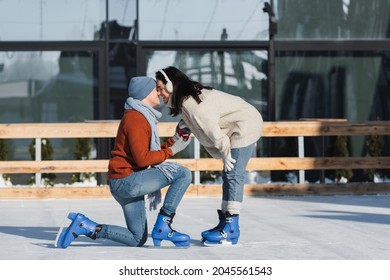 full length of young man holding box with wedding ring and kneeling while making proposal and kissing with woman on ice rink