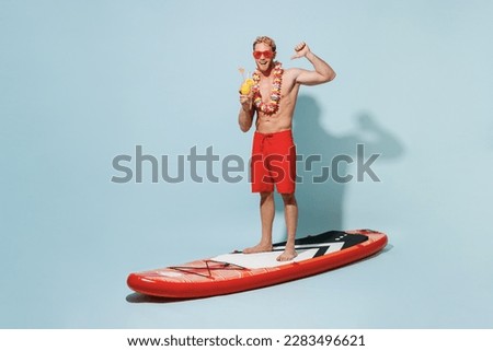 Full length young man in red shorts swimsuit hawaii lei glasses stand sup board hold alcohol cocktail point thumb finger on himself isolated on blue background Summer vacation sea rest sun tan concept