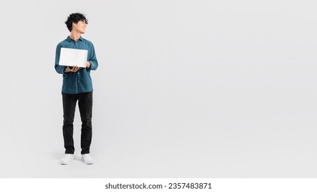 Full length of young man using laptop for work or studies, looking aside at free space while holding computer, advertising freelance online job or e-learning offer on white background. Panorama