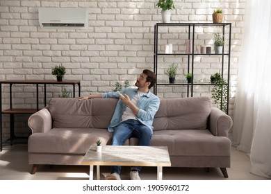 Full length young man wearing glasses holding using air conditioner remote controller, relaxing, sitting on couch, switching, setting comfort temperature in modern living room, enjoy fresh air