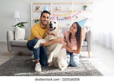 Full length of young multinational couple in party hats celebrating dog's birthday, posing and smiling at camera indoors. Affectionate owners having b-day party for their golden retriever at home - Powered by Shutterstock