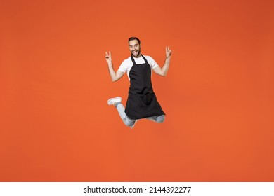 Full Length Young Overjoyed Man 20s Barista Bartender Barman Employee In Black Apron White T-shirt Work In Coffee Shop Jump High Isolated On Orange Background Studio . Small Business Startup Concept