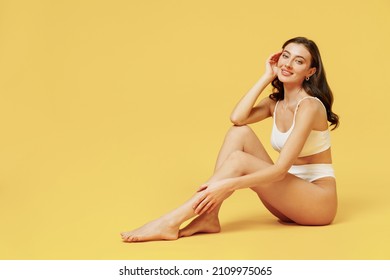 Full Length Young Sexy Brunette Woman 20s Wearing White Underwear With Perfect Fit Figure Sitting On Floor Touch Legs Soft Skin Isolated On Plain Yellow Color Background. People Female Beauty Concept.