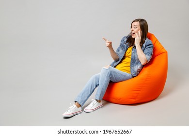 Full length young shocked confused caucasian woman 20s wearing casual denim jacket yellow t-shirt look camera sitting in bean bag chair point finger aside isolated on grey background studio portrait