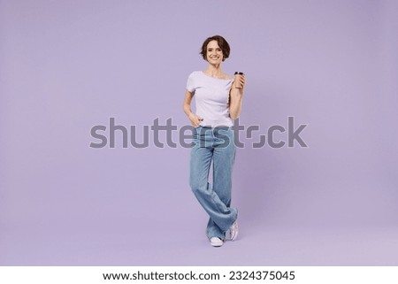 Full length young smiling cheerful fun happy woman 20s wear white t-shirt hold takeaway delivery craft paper brown cup coffee to go stand akimbo isolated on pastel purple background studio portrait
