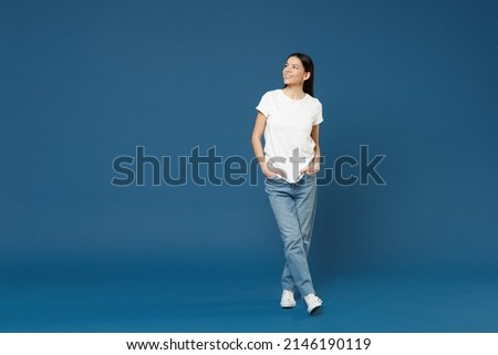 Full length of young smiling confident nice attractive beautiful latin woman 20s wearing white casual basic t-shirt keep hands in pockets look aside isolated on dark blue background studio portrait