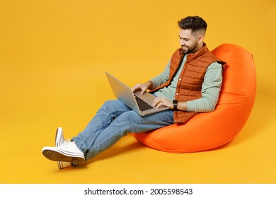 Full Length Young Smiling Freelancer Fun Caucasian Man 20s Years Old Wear Orange Vest Mint Sweatshirt Sitting In Beanbag Bag Chair Hold Laptop Pc Computer Isolated On Yellow Background Studio Portrait