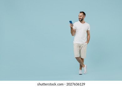 Full length young smiling friendly happy man 20s wearing casual white t-shirt looking camera using mobile cell phone chat onine isolated on plain pastel light blue color background studio portrait. - Shutterstock ID 2027636129