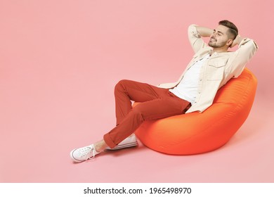 Full length young smiling happy overjoyed joyful fashionable man 20s in jacket white t-shirt sitting in bean bag chair resting hold hand behind neck isolated on pastel pink background studio portrait.