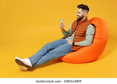 Full length young smiling happy fun caucasian man 20s years old wear orange vest mint sweatshirt sitting in beanbag bag chair hold mobile cell phone chat isolated on yellow background studio portrait