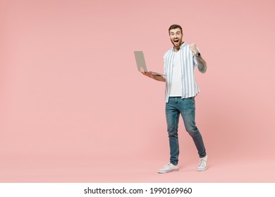 Full length young smiling happy fun unshaven man in blue striped shirt holding laptop pc computer chat online browsing internet do winner gesture clench fist isolated on pastel pink background studio.